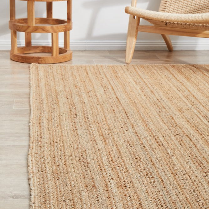 Beige woven rug on a wooden floor with a side table and chair in the background.
