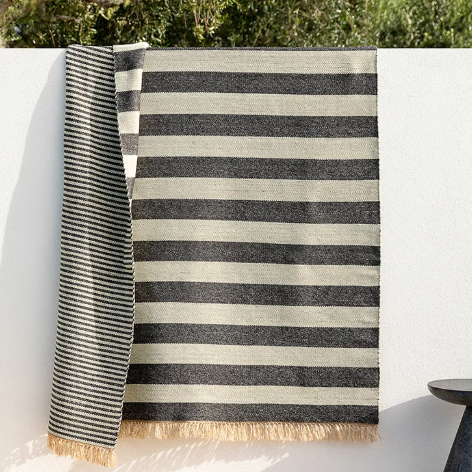 A striped woven throw blanket with cream and charcoal gray horizontal stripes and fringed edges, draped over a white wall outdoors with green foliage above.