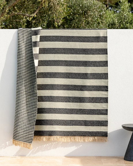 A striped woven throw blanket with cream and charcoal gray horizontal stripes and fringed edges, draped over a white wall outdoors with green foliage above.