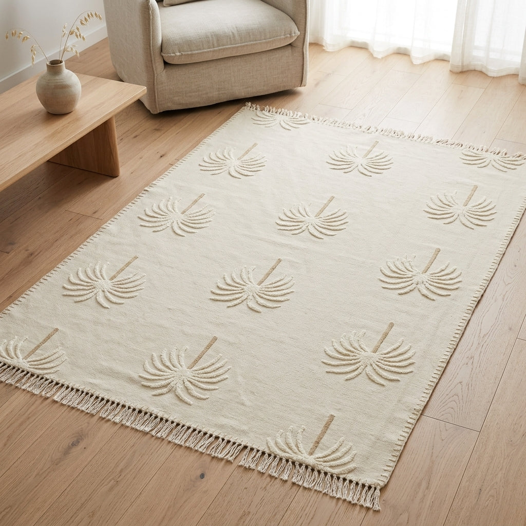 A cream-colored area rug with a raised palm leaf pattern and fringe, shown on a light wood floor in a bright living room next to a beige armchair and a wooden side table.