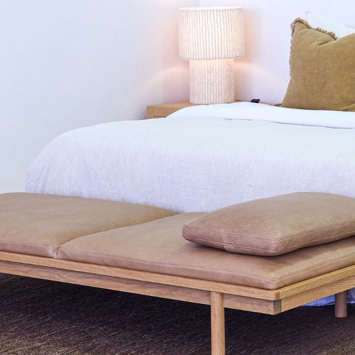Wooden bench with cushions in front of a bed with white bedding and a lamp.