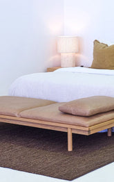 Wooden bench with cushions in front of a bed with white bedding and a lamp.