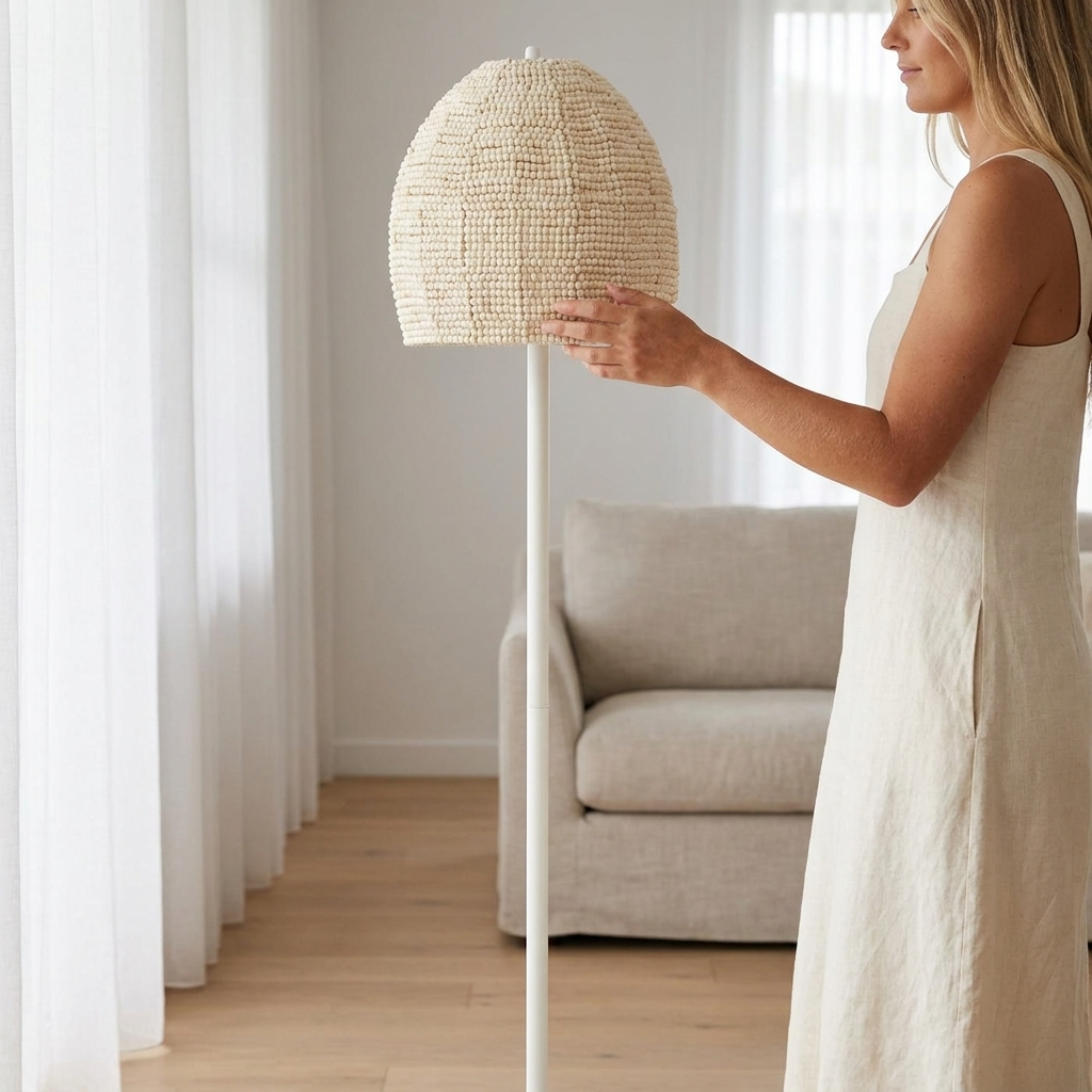 A woman in a white sleeveless dress stands in a bright room holding a tall floor lamp with a woven beige shade and a white pole.