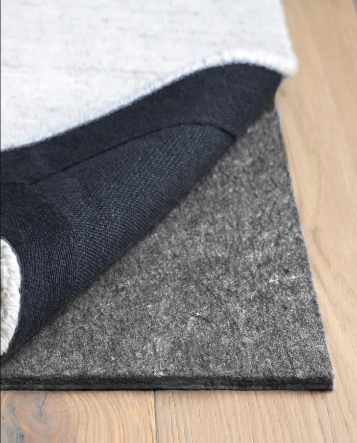A close-up of a dark gray felt floor mat with a black backing and a light gray top layer, shown partially peeled back on a light wood floor.