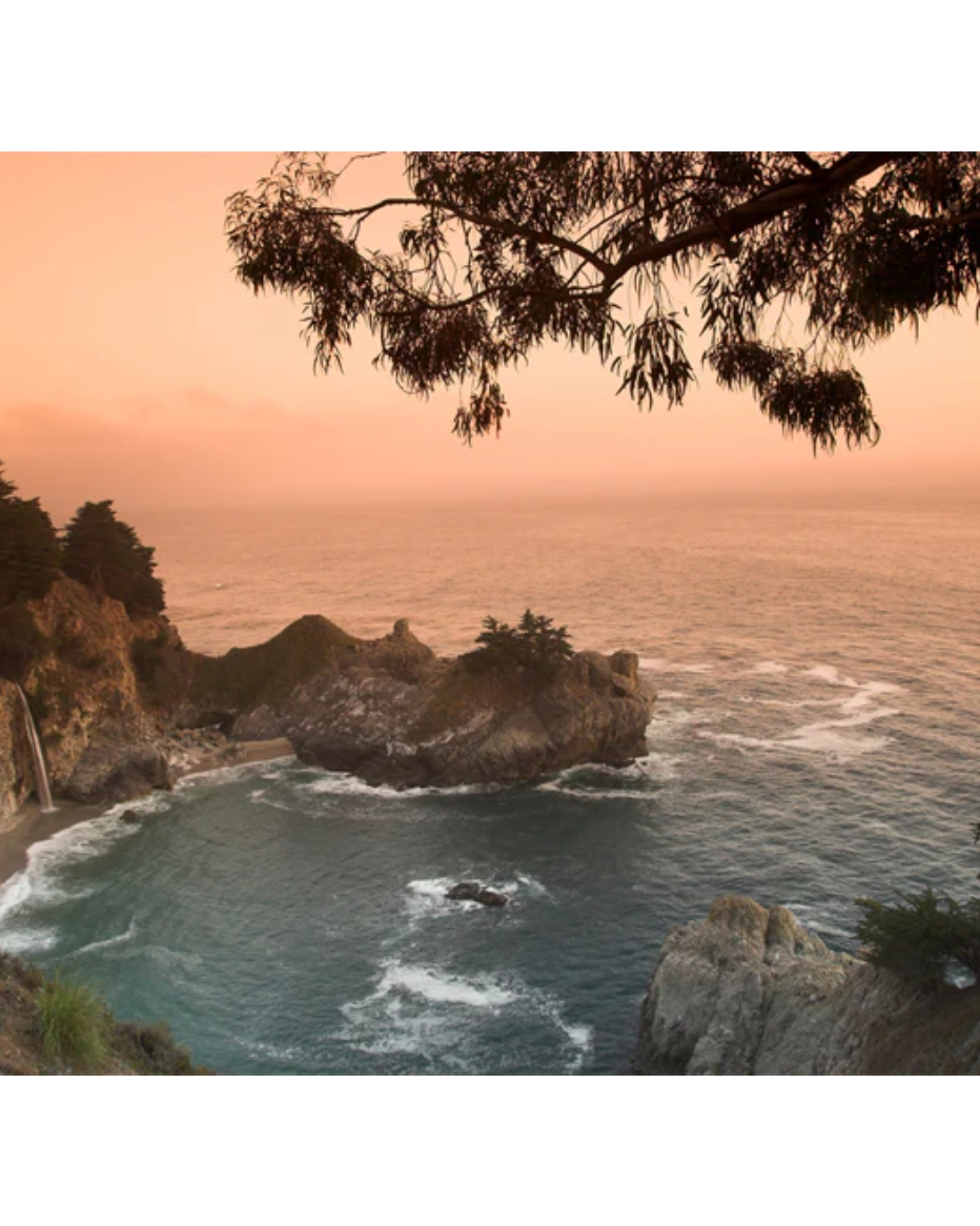 A scenic landscape photograph of a rocky coastal inlet at sunset, with orange sky, blue-green ocean water, dark cliffs, and tree silhouettes framing the top and right edges, shown on a white background.