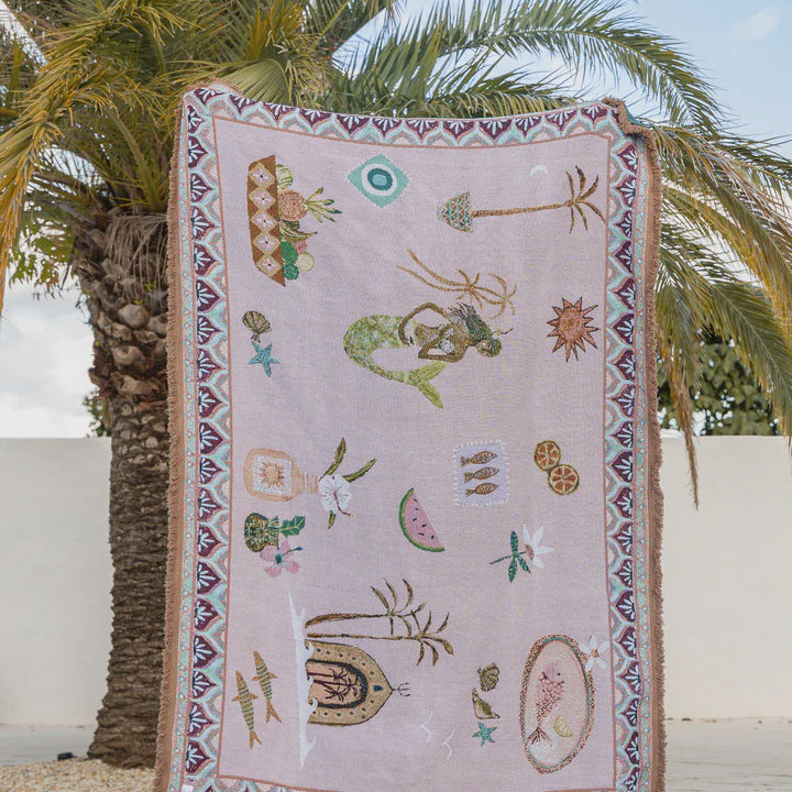 Decorative throw with mermaid and shell patterns hanging against a palm tree and white wall background.