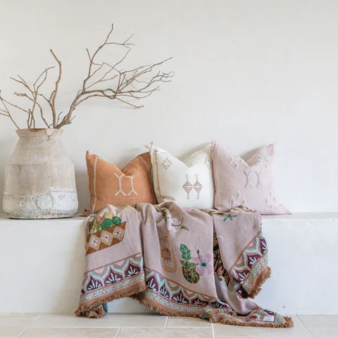 Decorative pillows and throw blanket on a bench with a vase and branches in the background.