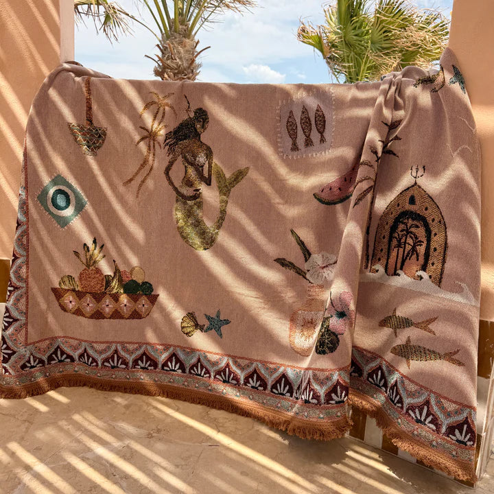 Decorative throw blanket with mermaid and fish designs draped over a chair outdoors.