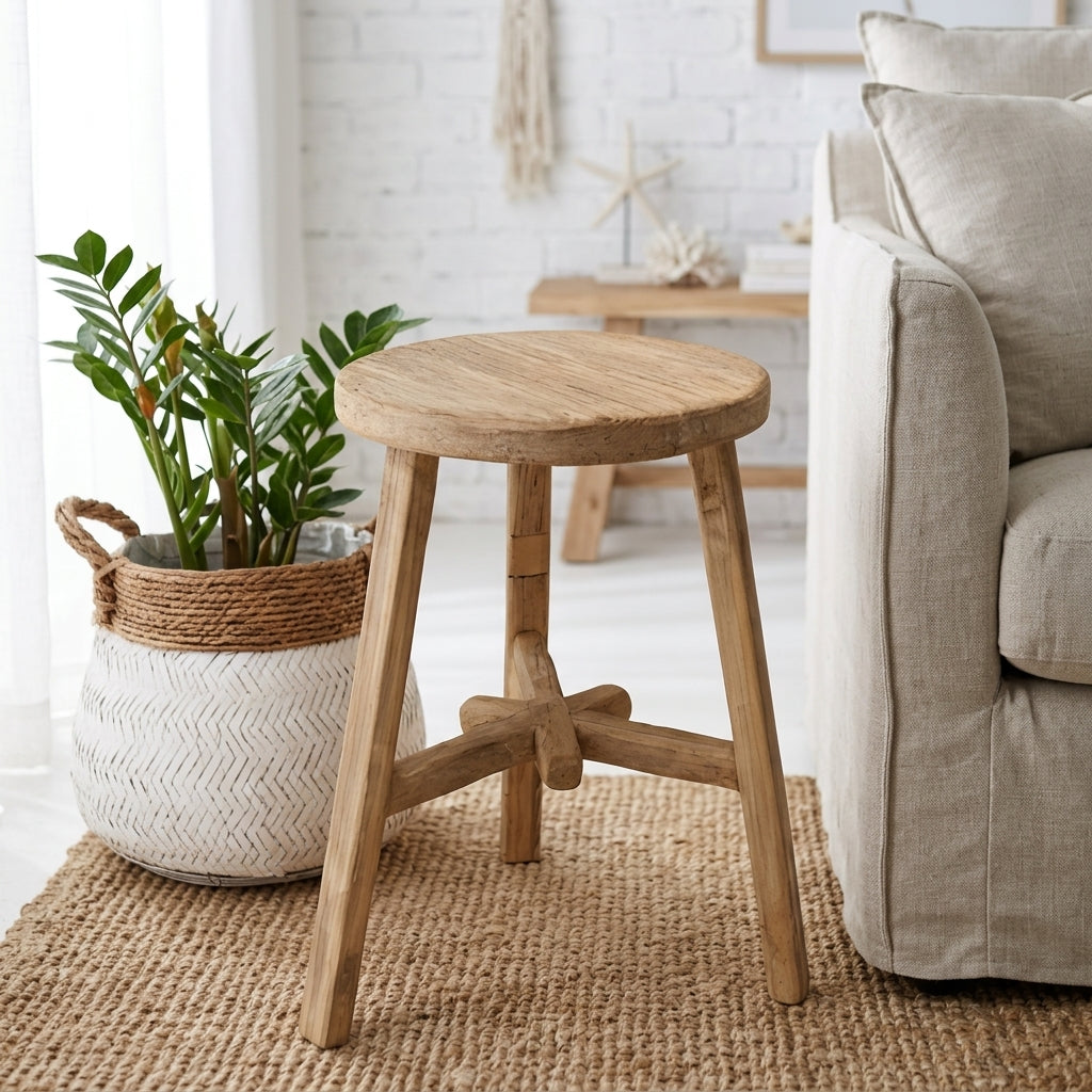 A round wooden stool with four legs and a central crossbar, shown in a bright living room next to a potted plant and a light beige sofa.