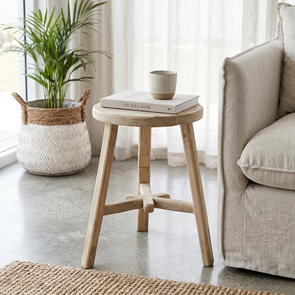 A small round wooden stool side table with three legs and a central crossbar, styled in a bright living room next to a beige armchair with a book and cup on top.