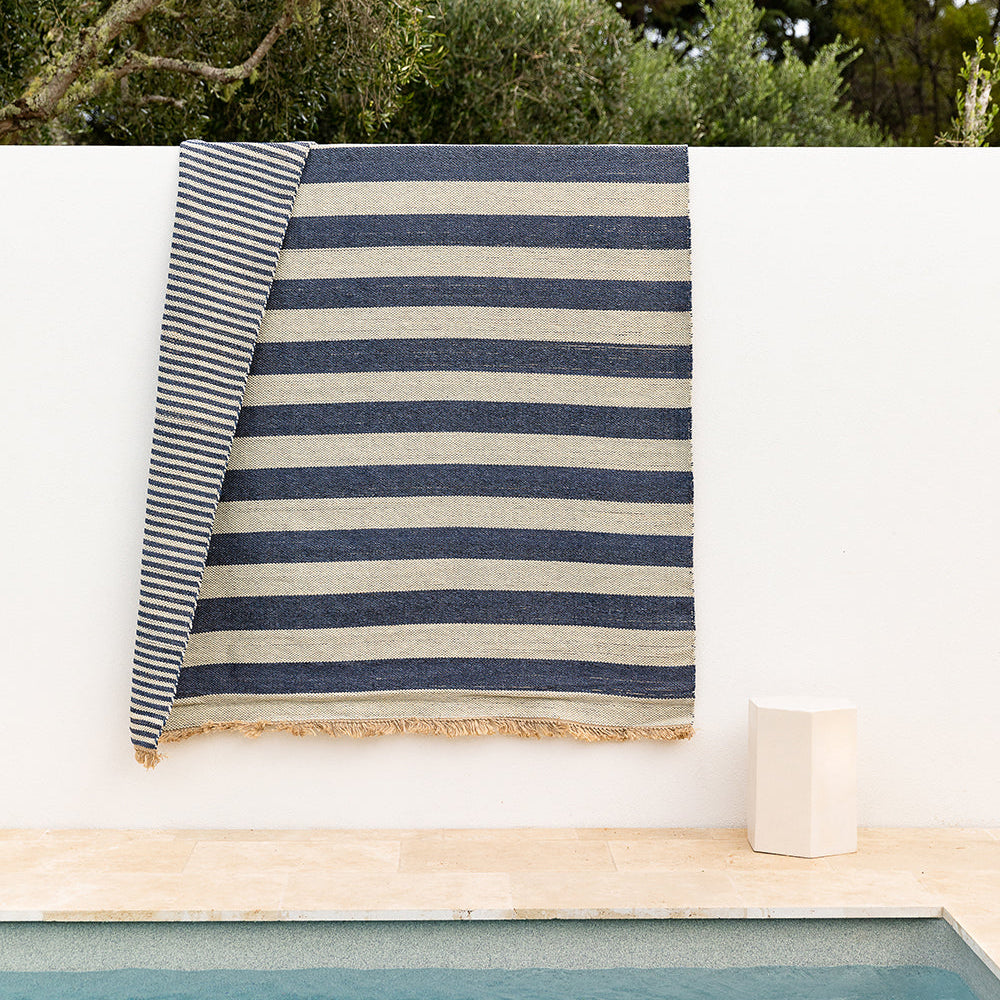 A striped woven textile, likely a beach towel or blanket, draped over a white wall above a pool. The fabric features alternating navy blue and beige horizontal stripes with a fringed edge, set against a background of green trees and a beige stone deck.