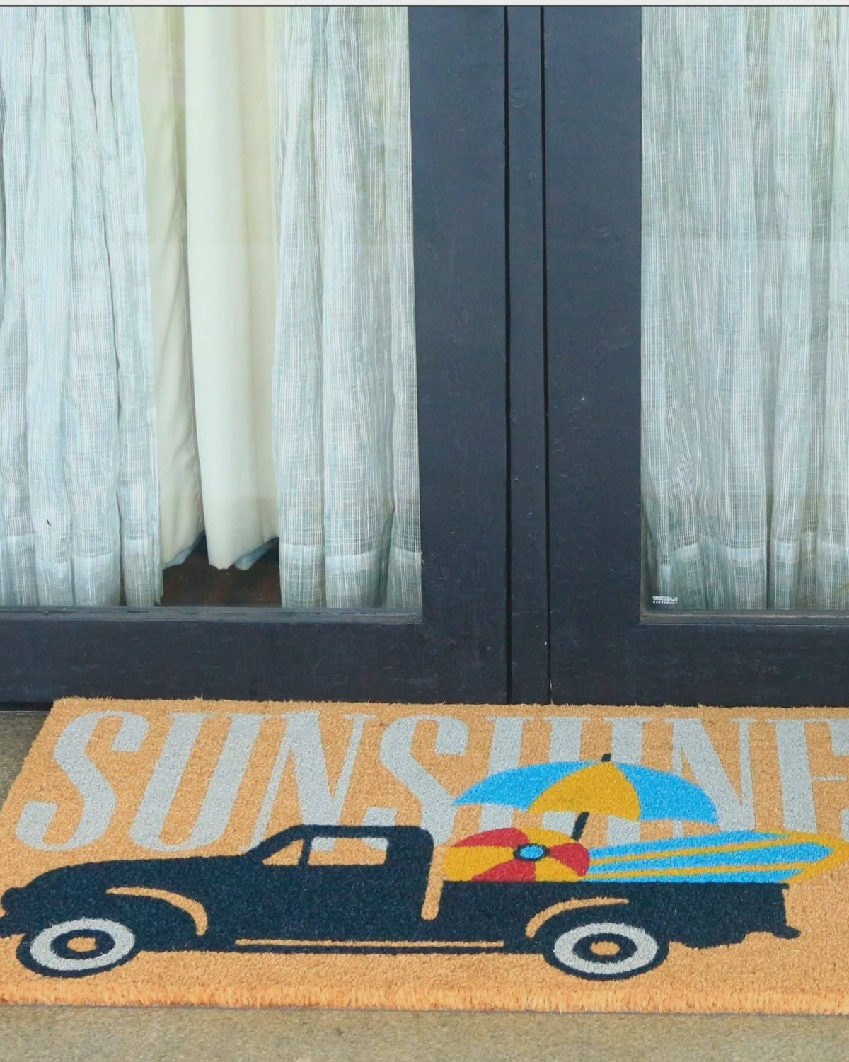 A rectangular coir doormat with a vintage pickup truck carrying surfboards and a beach ball, with the word 'SUNSHINE' in large letters across the top, placed in front of glass patio doors with curtains.