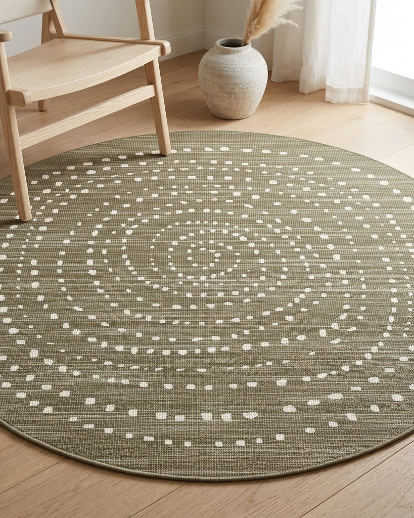A round area rug with a textured olive green base and a white dotted spiral pattern, styled on a light wood floor in a bright room next to a wooden chair and a vase.