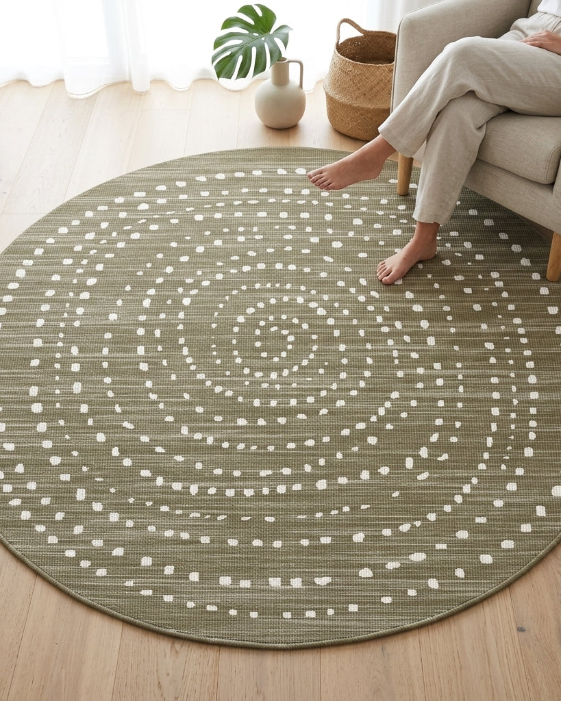 A round area rug with a textured olive green base and a white dotted spiral pattern, shown on a light wood floor in a living room setting with a person sitting on a beige armchair.