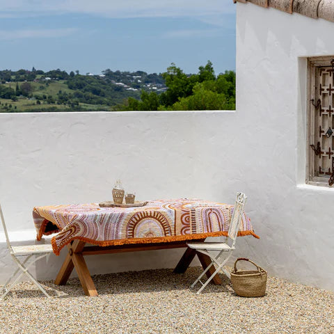 Outdoor setting with a table draped with a colorful table throw, chairs, and a woven basket against a white wall with a window.