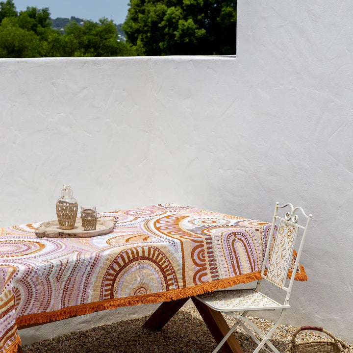 Outdoor setting with a table covered by a patterned tablecloth, chair, and basket against a white wall.
