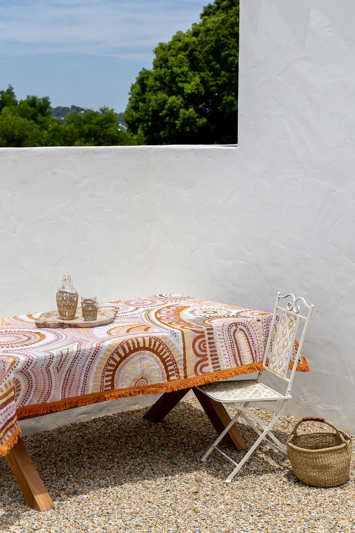 Outdoor setting with a table covered by a patterned tablecloth, chair, and basket against a white wall.