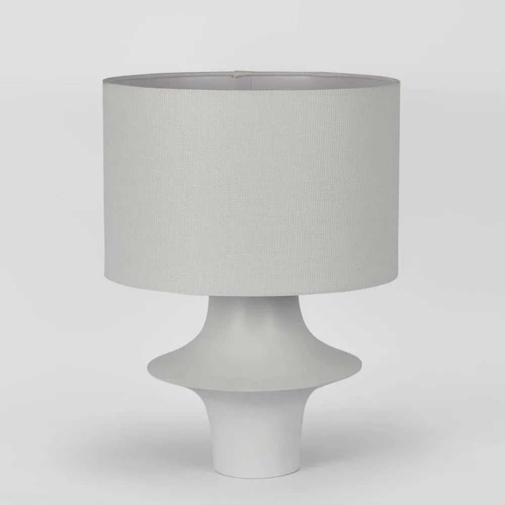 A modern table lamp with a textured light gray fabric drum shade and a sculptural, tiered white ceramic base, shown against a neutral light gray background.
