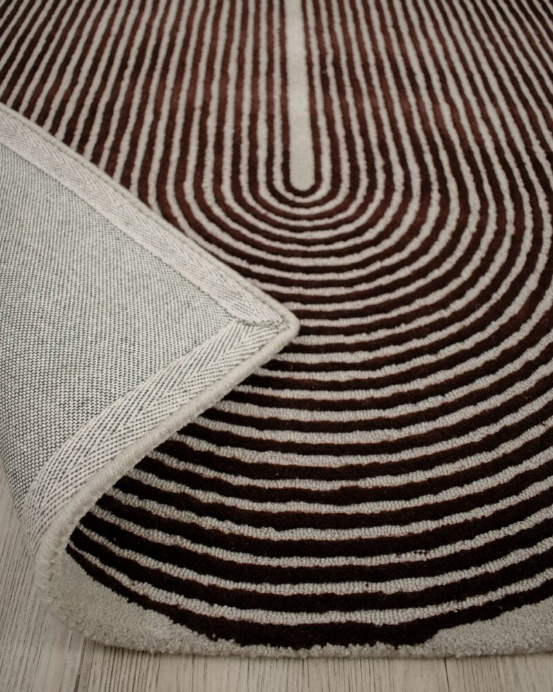 A close-up of a patterned area rug with concentric wavy stripes in dark brown and cream, with a folded corner showing the textured cream backing and edge.