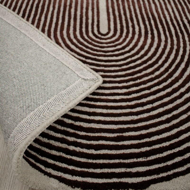 A close-up of a patterned area rug with concentric wavy stripes in dark brown and cream, with a folded corner showing the textured cream backing and edge.