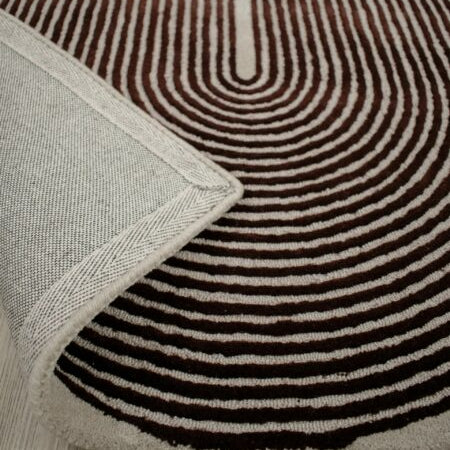 A close-up of a patterned area rug with concentric wavy stripes in dark brown and cream, with a folded corner showing the textured cream border, placed on a light wood floor.