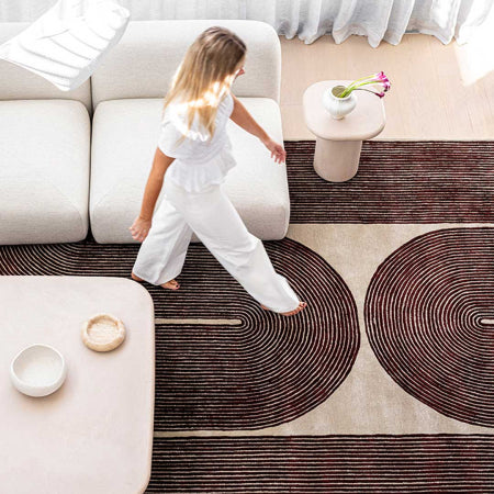A child in a white outfit walks barefoot across a large patterned area rug in a bright, modern living room with a white sofa, low tables, and sheer curtains.