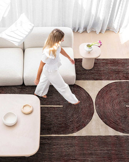 A child in a white outfit walks barefoot across a large patterned area rug in a bright, modern living room with a white sofa, low tables, and sheer curtains.