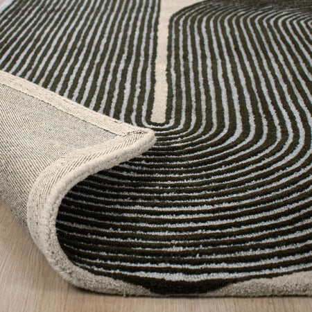 A close-up of a modern area rug with a dark charcoal and cream wavy geometric pattern, shown with one corner rolled up to reveal the backing and edge, placed on a light wood floor.