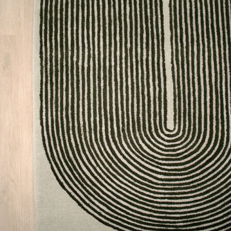 A close-up of a textured area rug with a bold black concentric arch pattern on a light beige background, shown on a light wood floor.