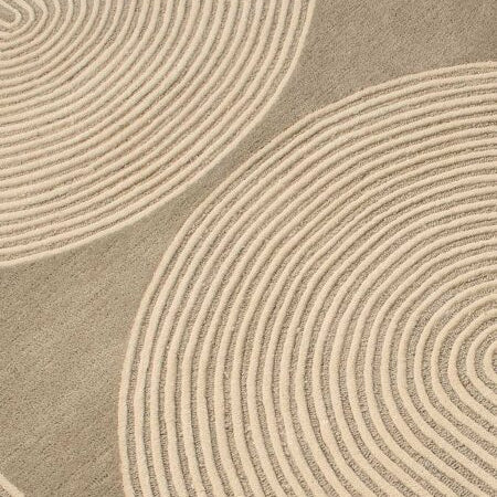 A close-up of a textured area rug featuring a modern abstract pattern of concentric wavy lines in warm beige and taupe-gray.