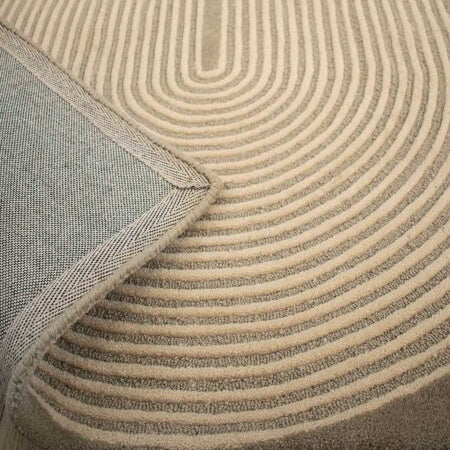A close-up of a textured area rug with concentric curved stripes in beige and taupe, showing the underside with a woven gray backing and a folded corner revealing the wood floor underneath.