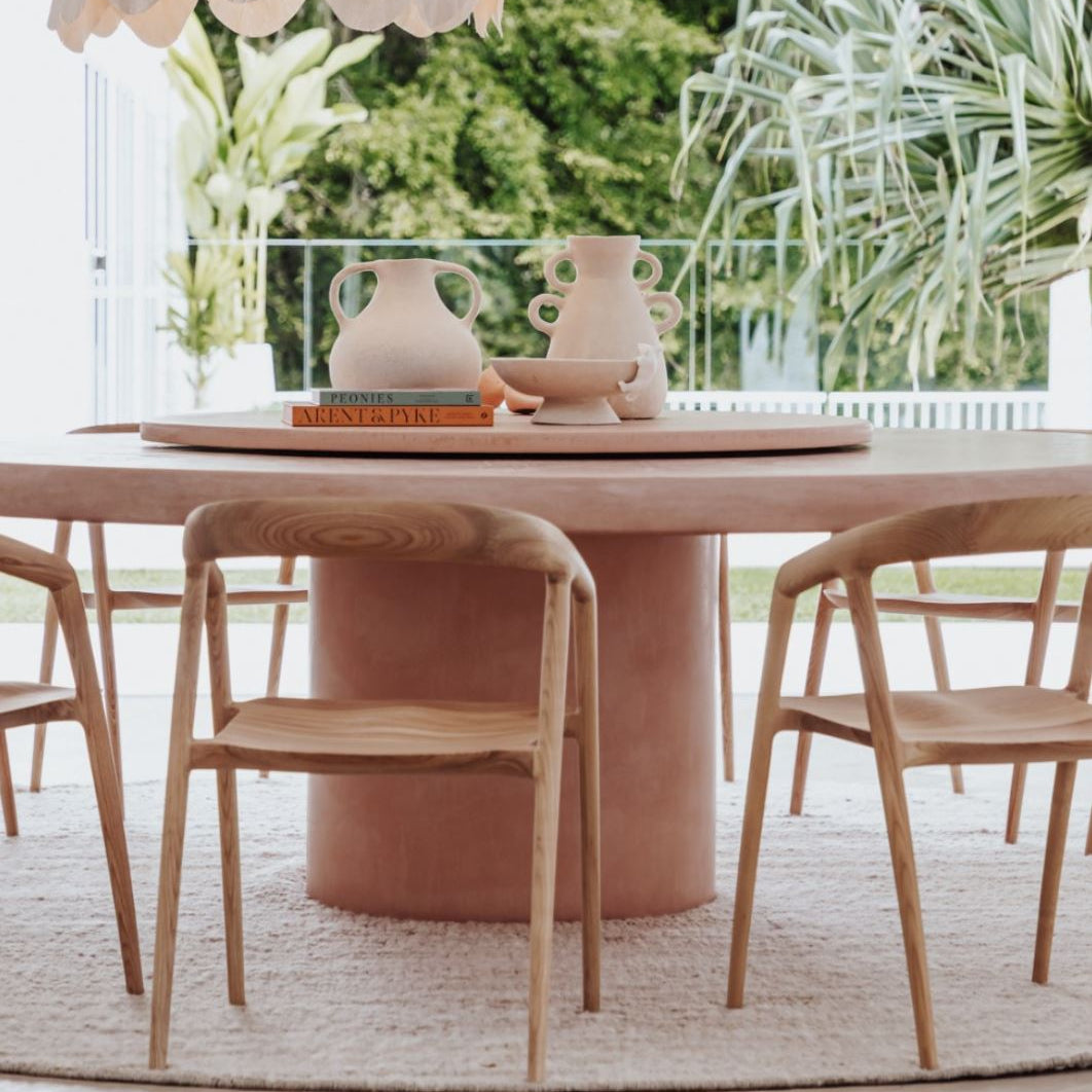 Dining area with a pink table and wooden chairs, surrounded by greenery.