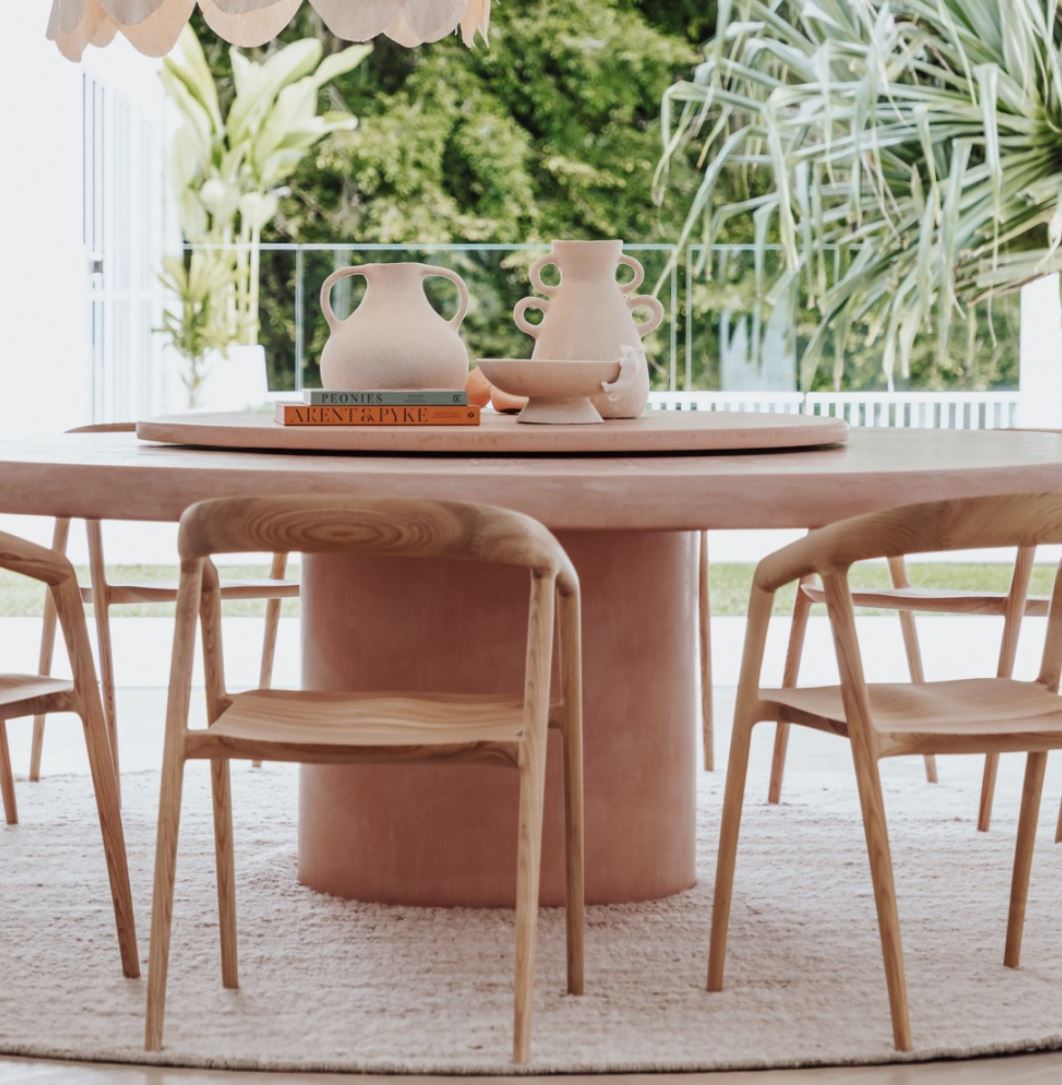 Dining area with a pink table and wooden chairs, surrounded by greenery.