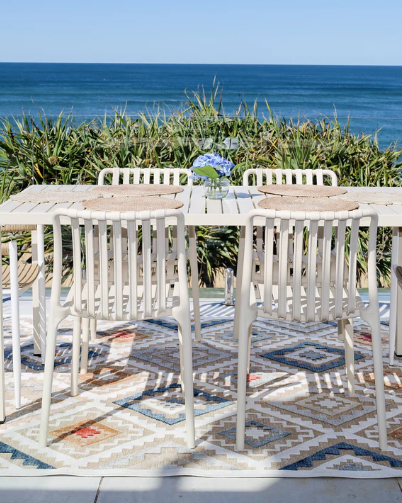 Outdoor dining set with white table and chairs on a patio by the ocean.