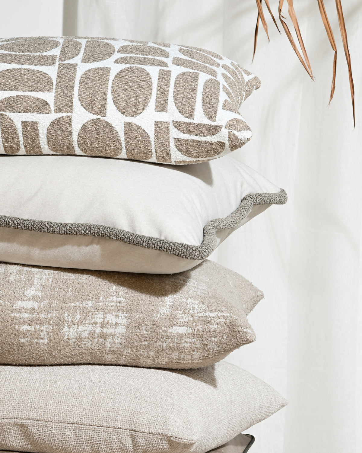 A stack of four decorative throw pillows in neutral beige and white tones, including a patterned pillow on top, set against a soft white curtain background with dried palm leaves in the corner.