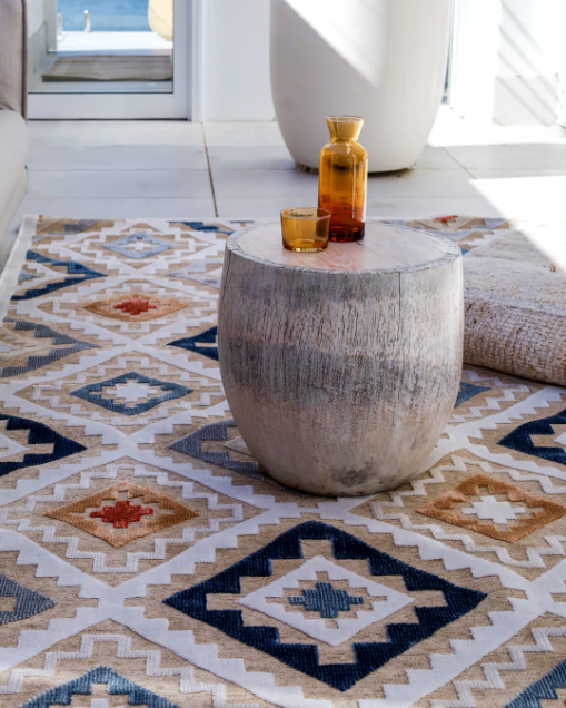 Decorative stone table with a glass and vase on a patterned rug in a bright room.