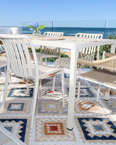 Outdoor dining set with table and chairs on a patterned rug by the sea.
