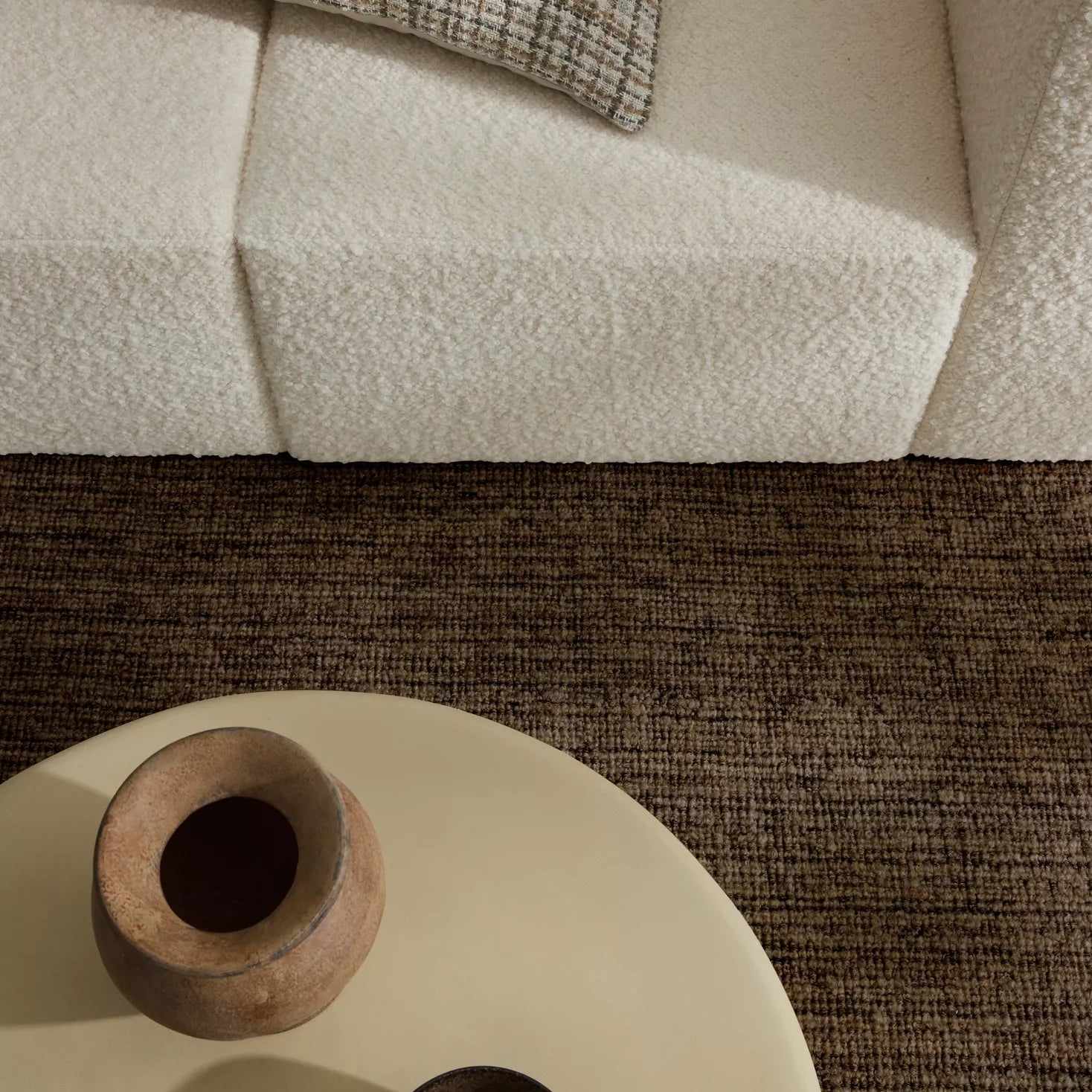 Round beige coffee table with wooden vase and bowl on a textured brown rug, next to a beige sofa with a patterned pillow.
