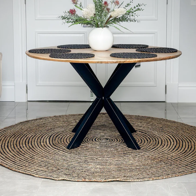 Round wooden table with black metal legs on a woven rug in a room.