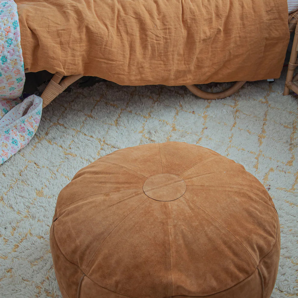 Brown ottoman on a textured floor with a bed and blanket in the background