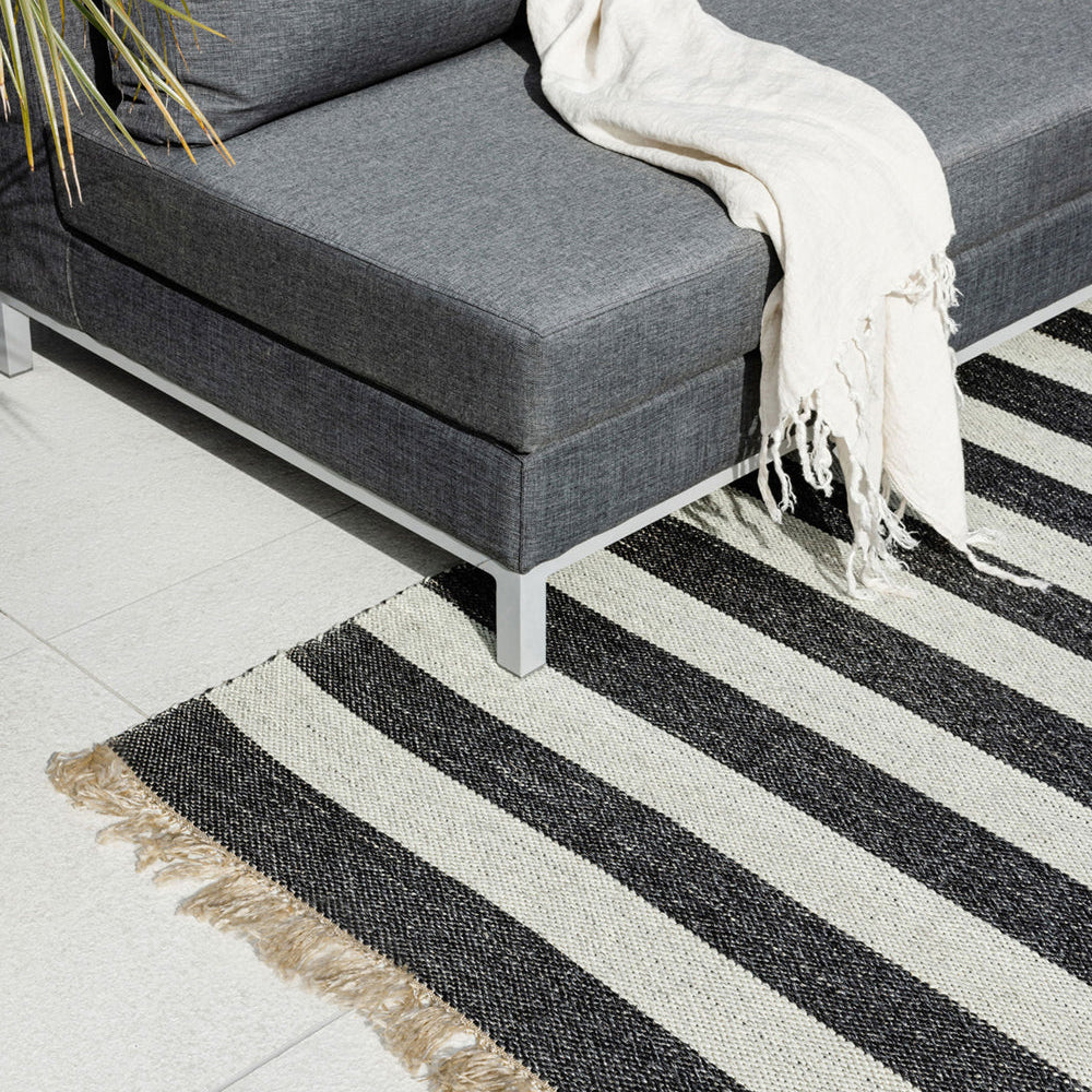 A modern gray sectional sofa with a white throw blanket, styled on a black and off-white striped woven area rug with fringed edges over a light tile floor.
