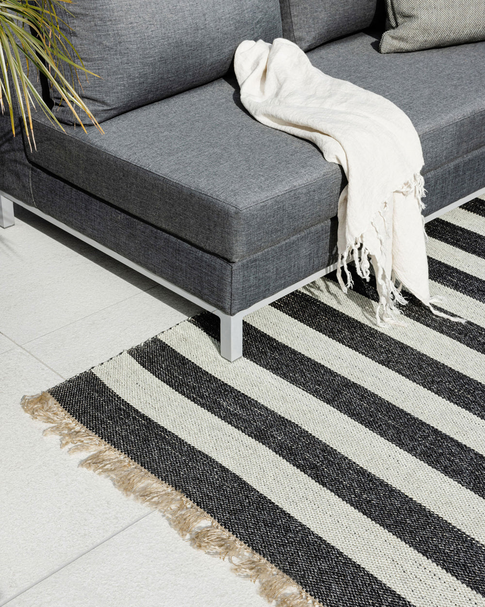 A modern gray sectional sofa with a white throw blanket, styled on a black and off-white striped woven area rug with fringed edges over a light tile floor.