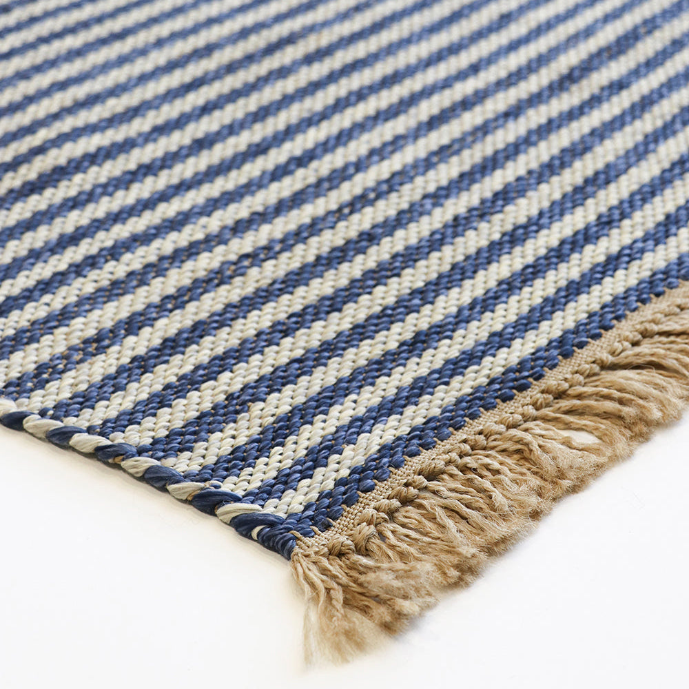 Close-up of a woven rug corner showing diagonal navy and cream stripes with a natural beige fringed edge on a white background.