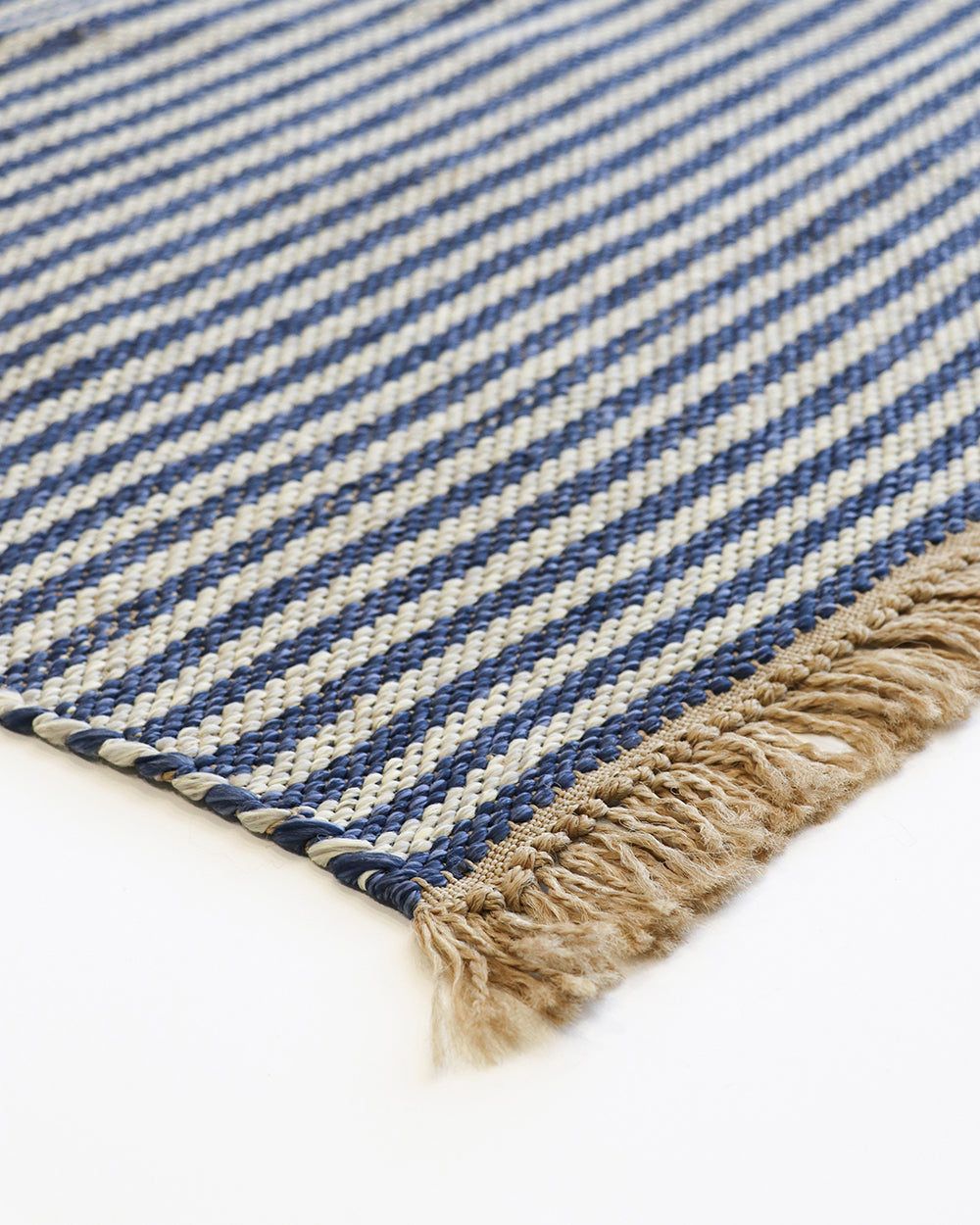 Close-up of a woven rug corner showing diagonal navy and cream stripes with a natural beige fringed edge on a white background.