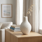 A ribbed ceramic vase in an off-white color, holding a dried decorative grass stem, styled on a light wood sideboard in a bright, modern living room.