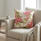 A square throw pillow with a pink floral and green leaf pattern on a cream background, styled on a beige armchair next to a small wooden side table with a mug.