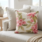 A square decorative throw pillow with a pink floral print on a cream background, styled on a light beige sofa next to a matching neutral pillow.
