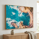 A large framed abstract coastal artwork hangs on a white wall above a light wood console table. The print shows vivid turquoise water, white surf, and warm tan/beige shoreline tones, surrounded by a thin gold-toned frame.