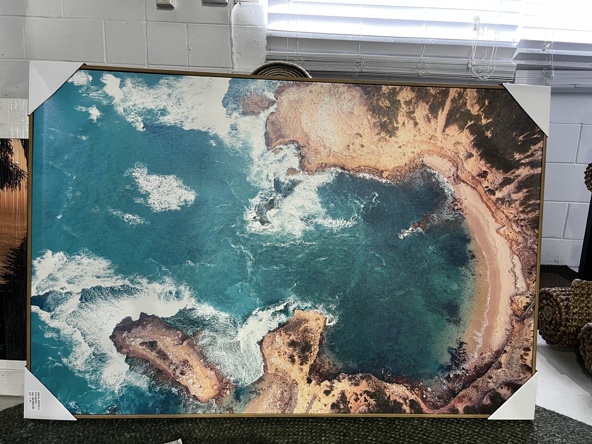 A large framed aerial photograph of a coastal bay with turquoise water, white surf, and sandy shoreline, displayed indoors with corner protectors on a gallery-wrapped canvas.