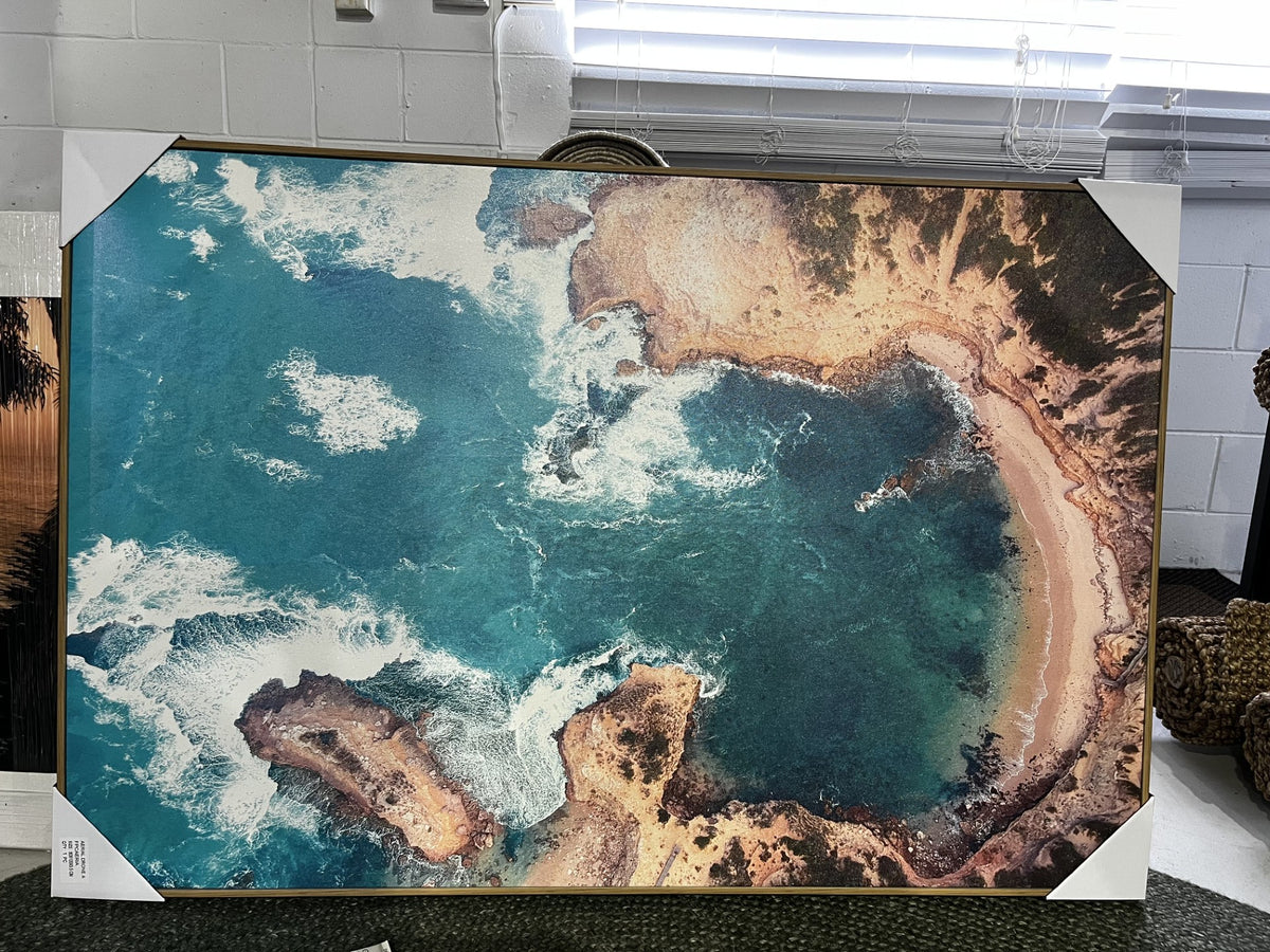 A large framed aerial photograph of a coastal bay with turquoise water, white surf, and sandy shoreline, displayed indoors with corner protectors on a gallery-wrapped canvas.